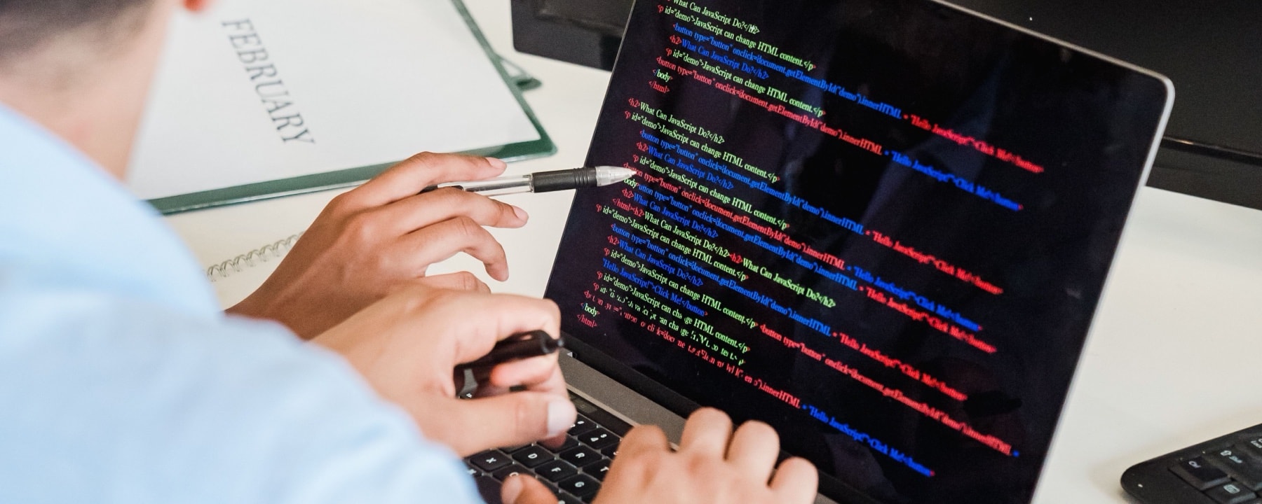 Persona trabajando en una notebook con código en pantalla, con una libreta y una lapicera sobre el escritorio.