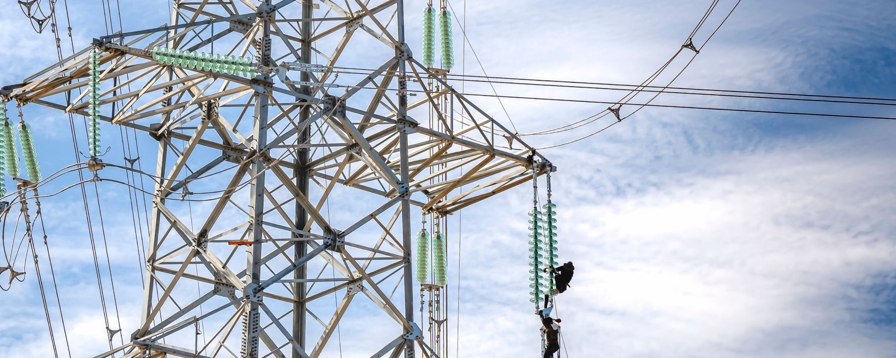 Dos operarios trabajando sobre el brazo de una torre de alta tensión, entre cadenas de aisladores, con el cielo de fondo.