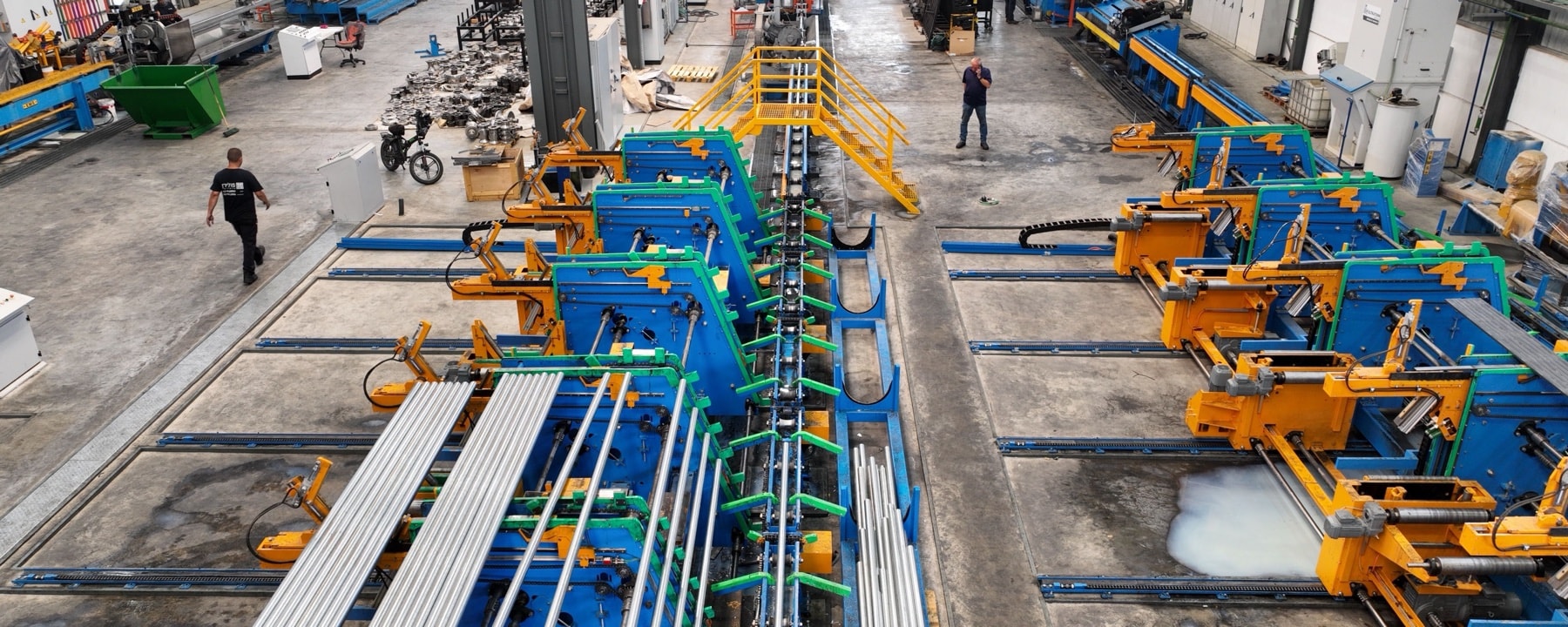 Nave industrial vista desde arriba, con dos líneas de producción de perfiles metálicos y operarios trabajando entre ellas.