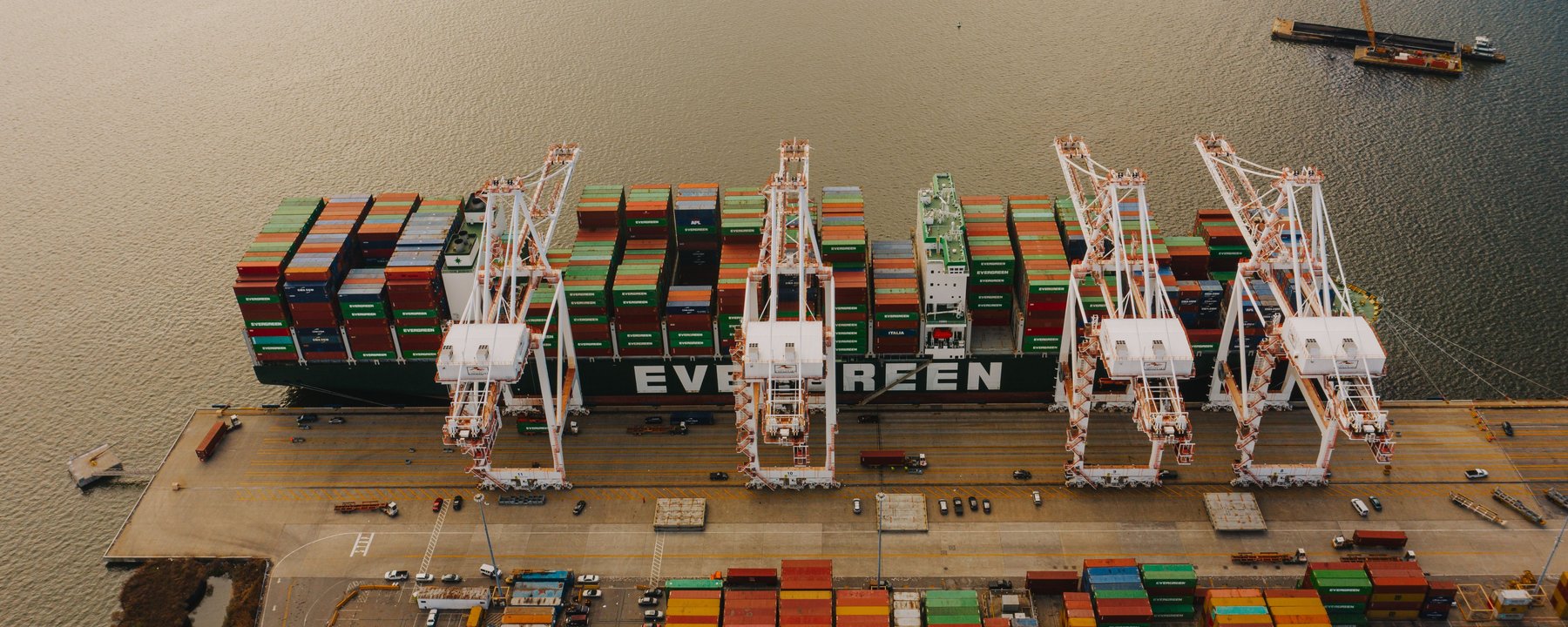 Vista aérea de un portacontenedores amarrado a muelle, con cuatro grúas pórtico y contenedores apilados en la explanada.